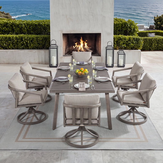 An outdoor patio dining set arranged around a rectangular gray table with six swivel armchairs, set on a light rug in front of a fireplace and ocean view. The table and chairs have light beige cushions, with the chairs featuring gray metal swivel bases.