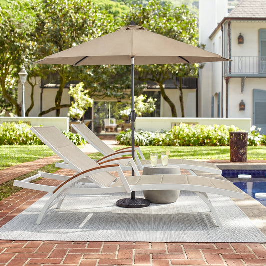 Beige patio umbrella over lounge chairs on a patio with a pool and garden in the background.