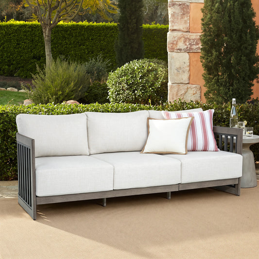 Outdoor patio sofa with light beige cushions and a dark gray frame, styled with two accent pillows in a garden setting.