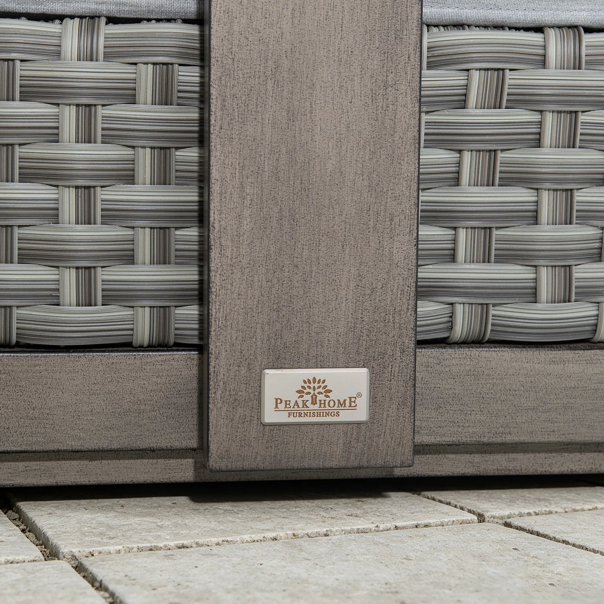 Extreme close-up of the Peak Home Furnishings metal logo plate on the weathered-finish frame next to the hand-woven gray PE wicker weave.