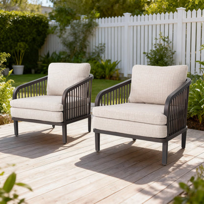 Two outdoor chairs with beige cushions on a wooden deck, surrounded by greenery and a white fence.