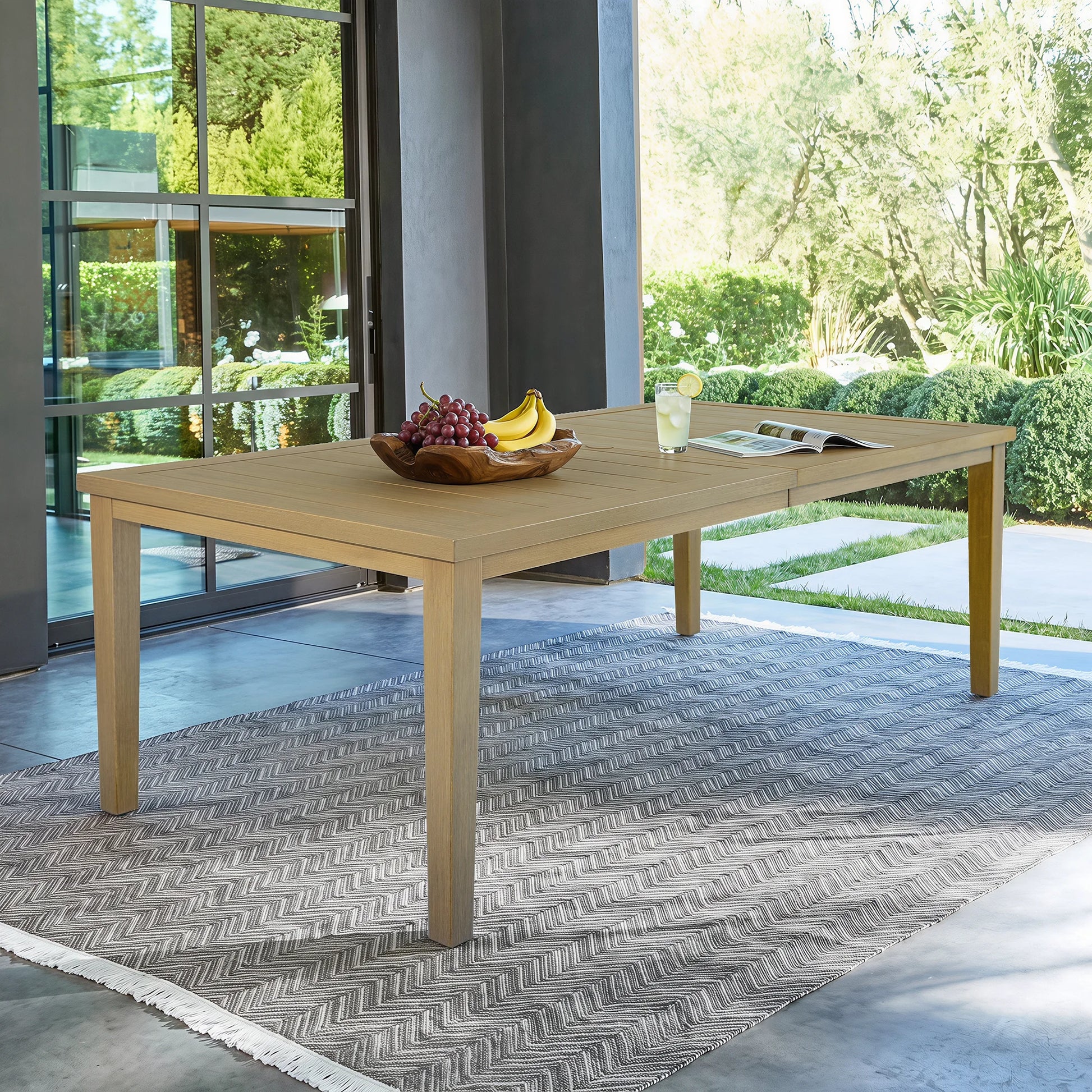 The Alcudia wood-look aluminum patio dining table styled on a grey chevron outdoor rug under a modern covered porch.