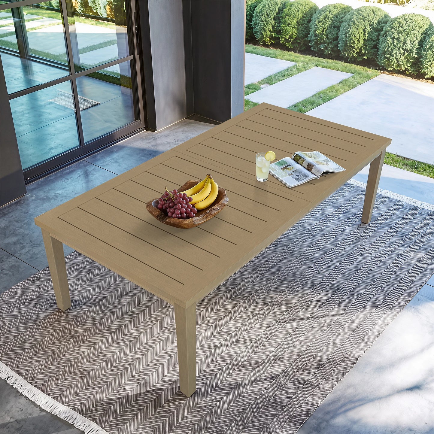High-angle overhead view of the rectangular Alcudia outdoor dining table placed on a patterned patio rug next to a green lawn.