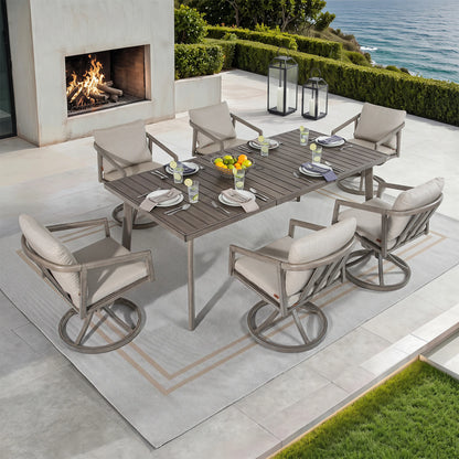 A 7-piece outdoor patio dining set with a rectangular slat-top table and six swivel armchairs with light beige cushions, arranged on a patterned rug beside a fireplace and overlooking the ocean.