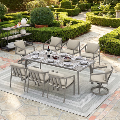 An 9-piece outdoor patio dining set with a rectangular metal table and six cushioned chairs, arranged on a striped area rug in a landscaped garden patio setting.