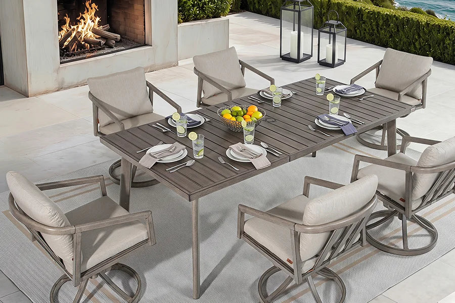A 7-piece outdoor patio dining set featuring a rectangular slat-top table and six swivel armchairs with beige cushions and gray metal frames, styled on a patio with a fireplace in the background.