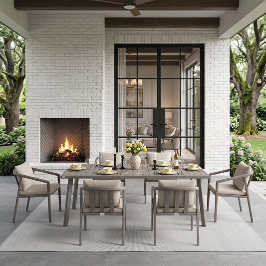 A 7-piece outdoor dining set with a rectangular weathered gray table and six matching slatted chairs with beige cushions, styled on a patio in front of a white brick fireplace and glass doors.