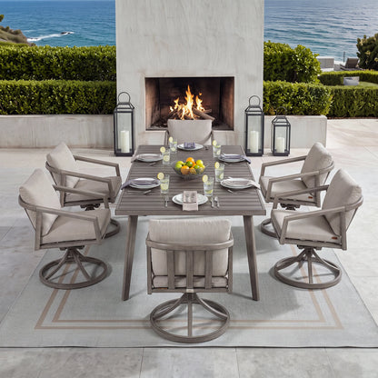 An outdoor patio dining set arranged around a rectangular gray table with six swivel armchairs, set on a light rug in front of a fireplace and ocean view. The table and chairs have light beige cushions, with the chairs featuring gray metal swivel bases.