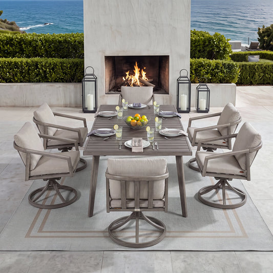 An outdoor patio dining set arranged around a rectangular gray table with six swivel armchairs, set on a light rug in front of a fireplace and ocean view. The table and chairs have light beige cushions, with the chairs featuring gray metal swivel bases.