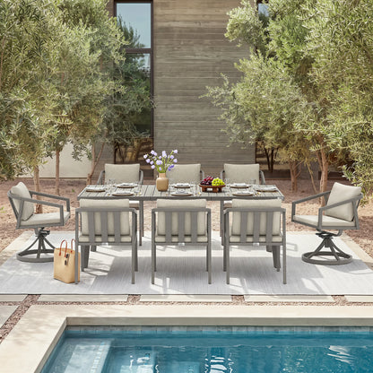 An outdoor dining set arranged on a light gray patio rug beside a swimming pool, with a modern wooden building and olive trees in the background. The set includes a rectangular dining table and six swivel armchairs with light gray cushions.