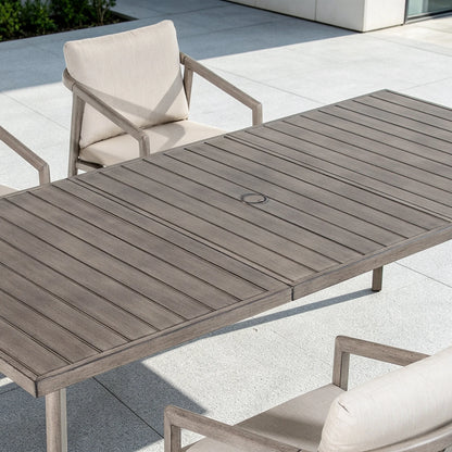 An outdoor rectangular patio dining table with weathered gray-brown slatted decking, shown with matching armchairs on a light stone patio.