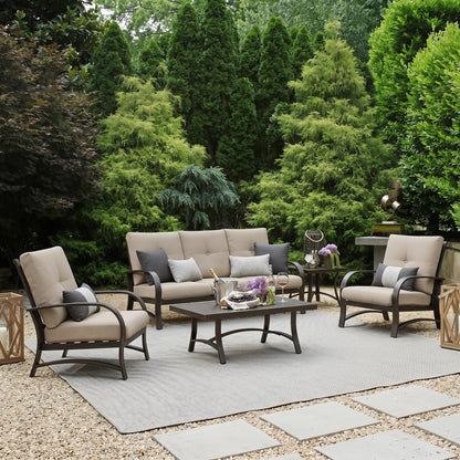 Outdoor patio set with beige sofa, chairs, and coffee table on a gravel patio with greenery in the background.