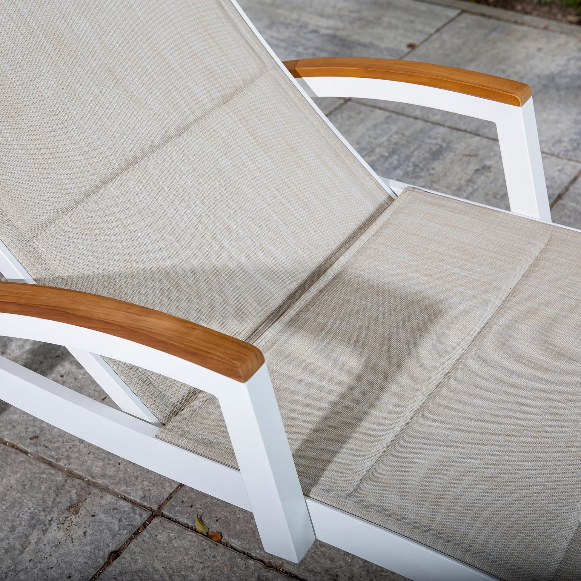 White lounge chair with beige cushions and wooden armrests on a stone patio.