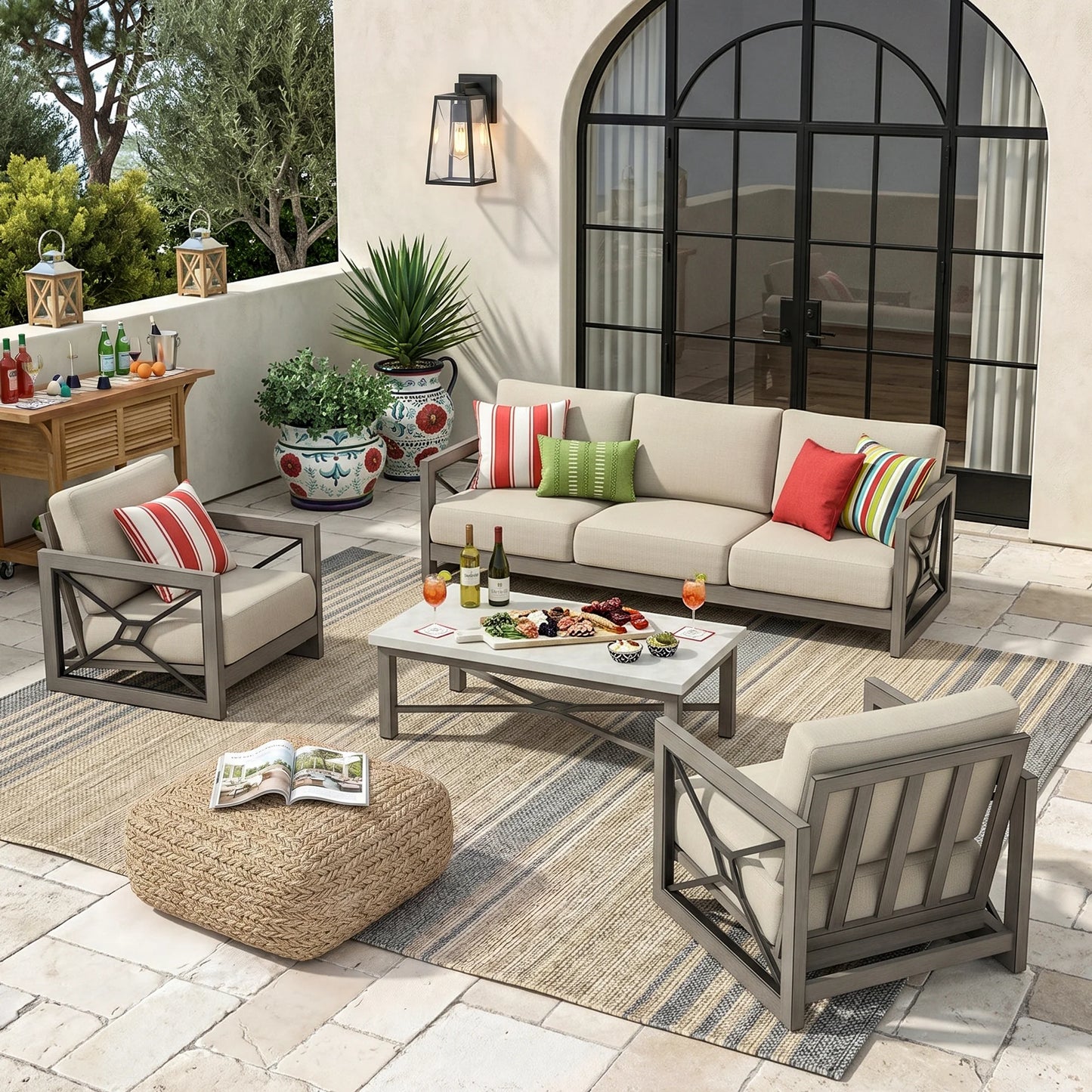 Outdoor patio furniture set arranged on a stone patio in front of a large arched glass door. The set includes a sofa, two armchairs, a rectangular coffee table, a woven pouf ottoman, and striped throw pillows in neutral beige, red, green, and multicolor tones.