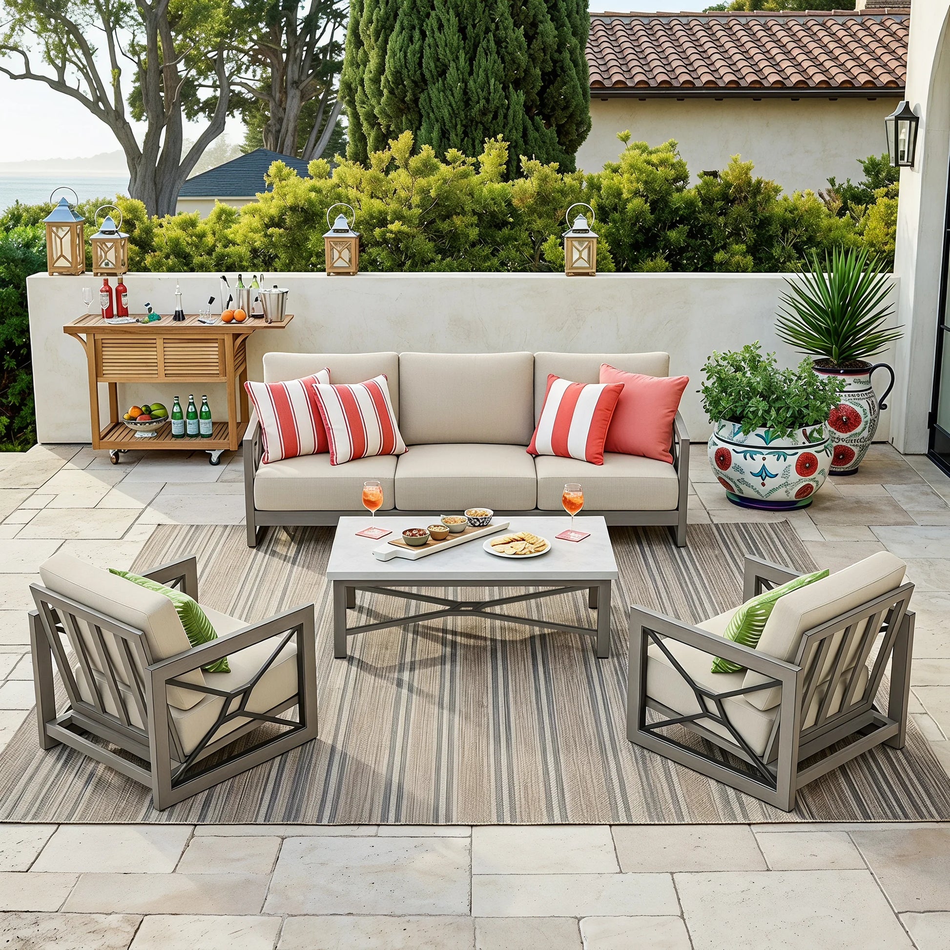 Outdoor patio furniture set including a beige three-seat sofa, two gray armchairs with cream cushions, a white coffee table, and a striped area rug, arranged on a stone patio with a bar cart and potted plants in the background.