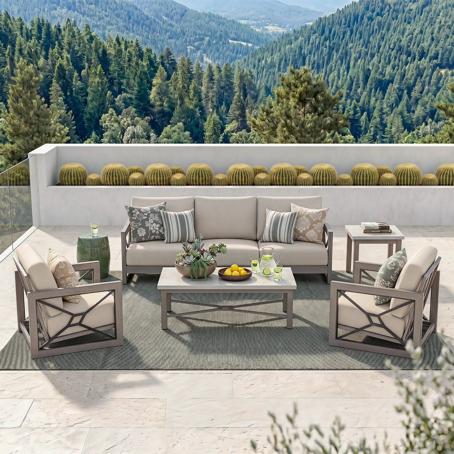 Outdoor patio furniture set arranged on a light stone terrace with a forested mountain backdrop. The set includes a beige sofa, two matching armchairs, a small side table, and a rectangular coffee table on a gray rug, with neutral cushions and decorative pillows.