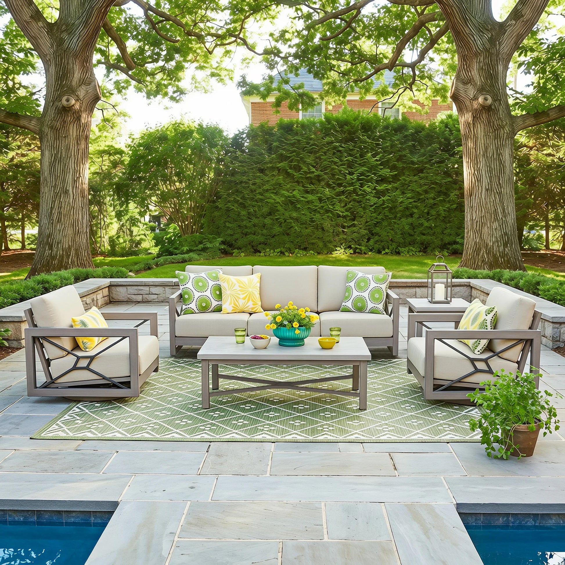 Outdoor patio furniture set arranged on a stone patio beside a pool, featuring a light beige sectional sofa, two matching armchairs, a rectangular coffee table, and a green patterned area rug. Large trees, a hedge, and landscaping are visible in the background.