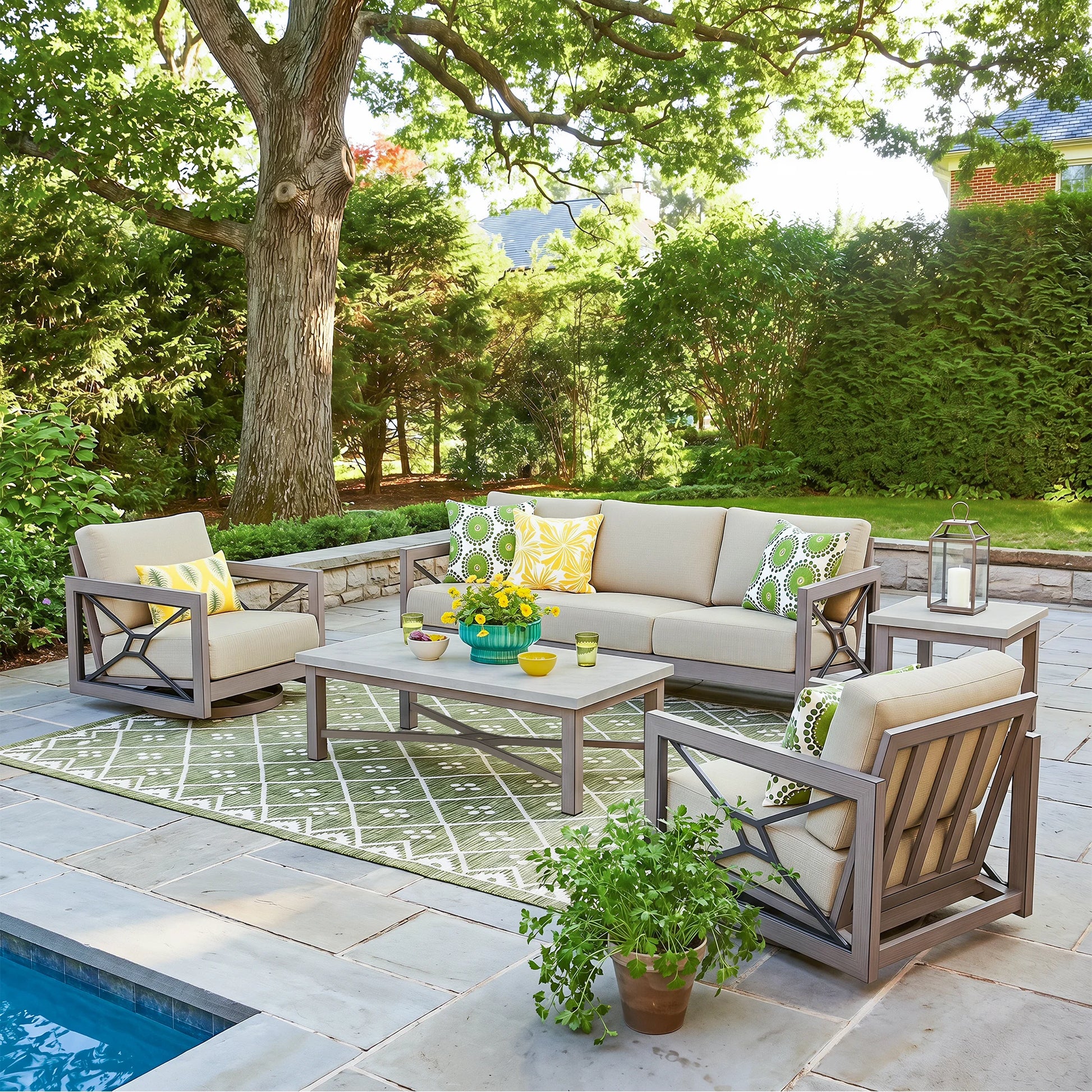 A 5-piece outdoor patio furniture set arranged on a stone patio next to a swimming pool. The set includes a sofa, two armchairs, a coffee table, and a side table, all in a gray frame with beige cushions and yellow and green patterned accent pillows.