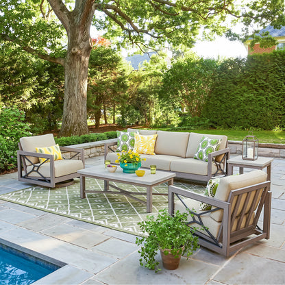 A 5-piece outdoor patio furniture set arranged on a stone patio next to a swimming pool. The set includes a sofa, two armchairs, a coffee table, and a side table, all in a gray frame with beige cushions and yellow and green patterned accent pillows.