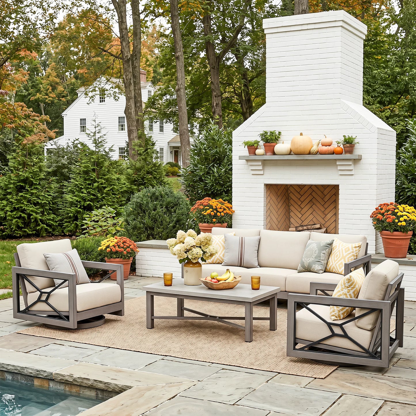 Outdoor patio furniture set arranged on a stone patio in front of a white brick fireplace, including two gray-framed armchairs with cream cushions, a matching sofa, a coffee table, and decorative pillows. Trees, flowers, and a house are visible in the background.