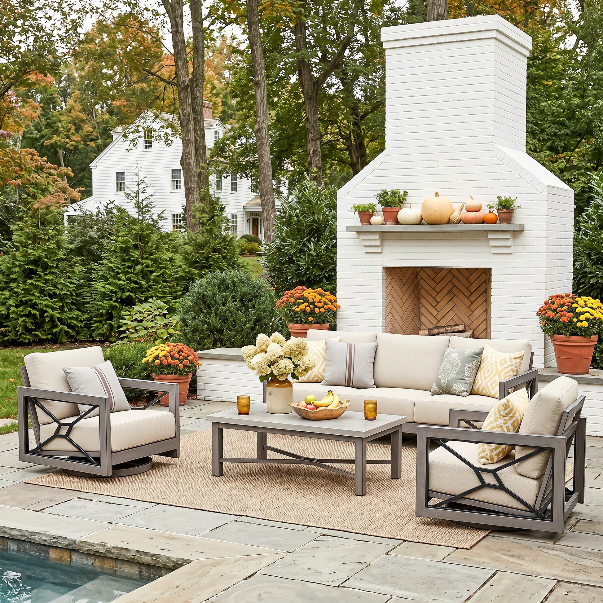 Outdoor patio furniture set arranged on a stone patio in front of a white brick fireplace, including two gray-framed armchairs with cream cushions, a matching sofa, a coffee table, and decorative pillows. Trees, flowers, and a house are visible in the background.