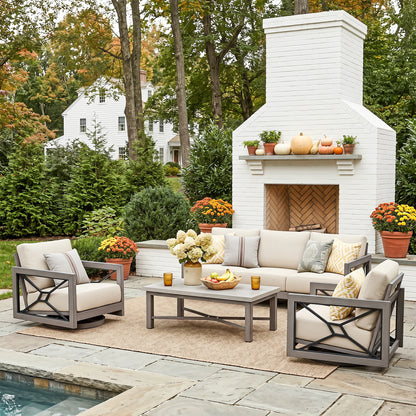 Outdoor patio furniture set arranged on a stone patio in front of a white brick fireplace, including two gray-framed armchairs with cream cushions, a matching sofa, a coffee table, and decorative pillows. Trees, flowers, and a house are visible in the background.