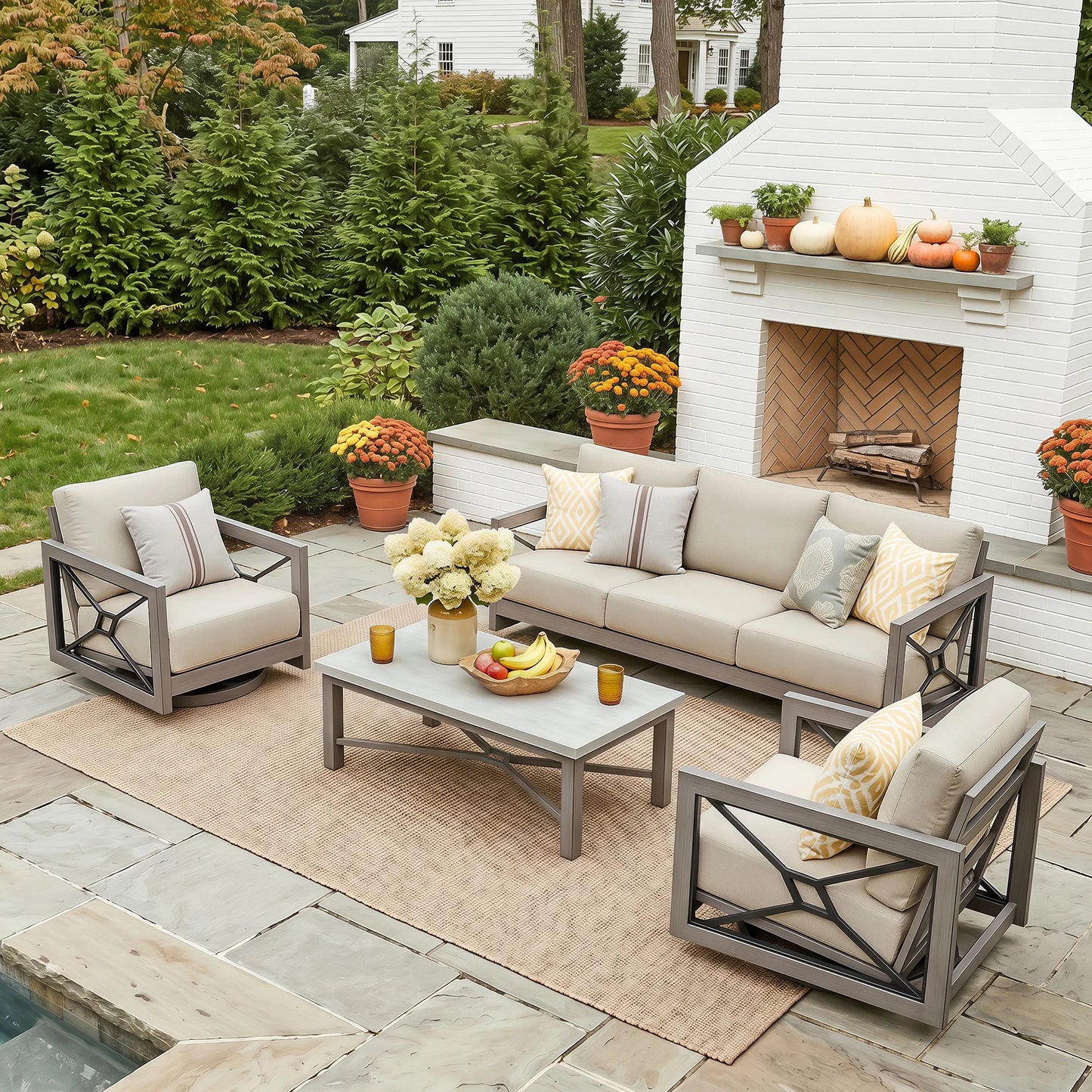 A 4-piece outdoor patio furniture set including a sofa, two armchairs, and a coffee table with neutral beige cushions and gray metal frames, arranged on a beige rug in front of a white brick fireplace.
