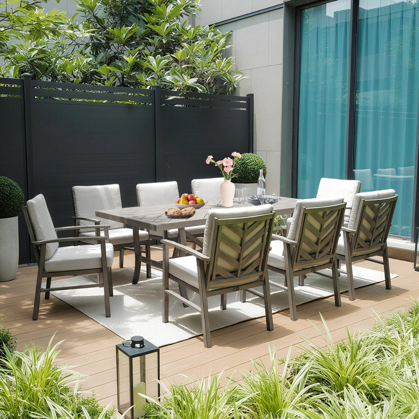 Outdoor dining set with table and chairs on a wooden deck, surrounded by plants and a building.