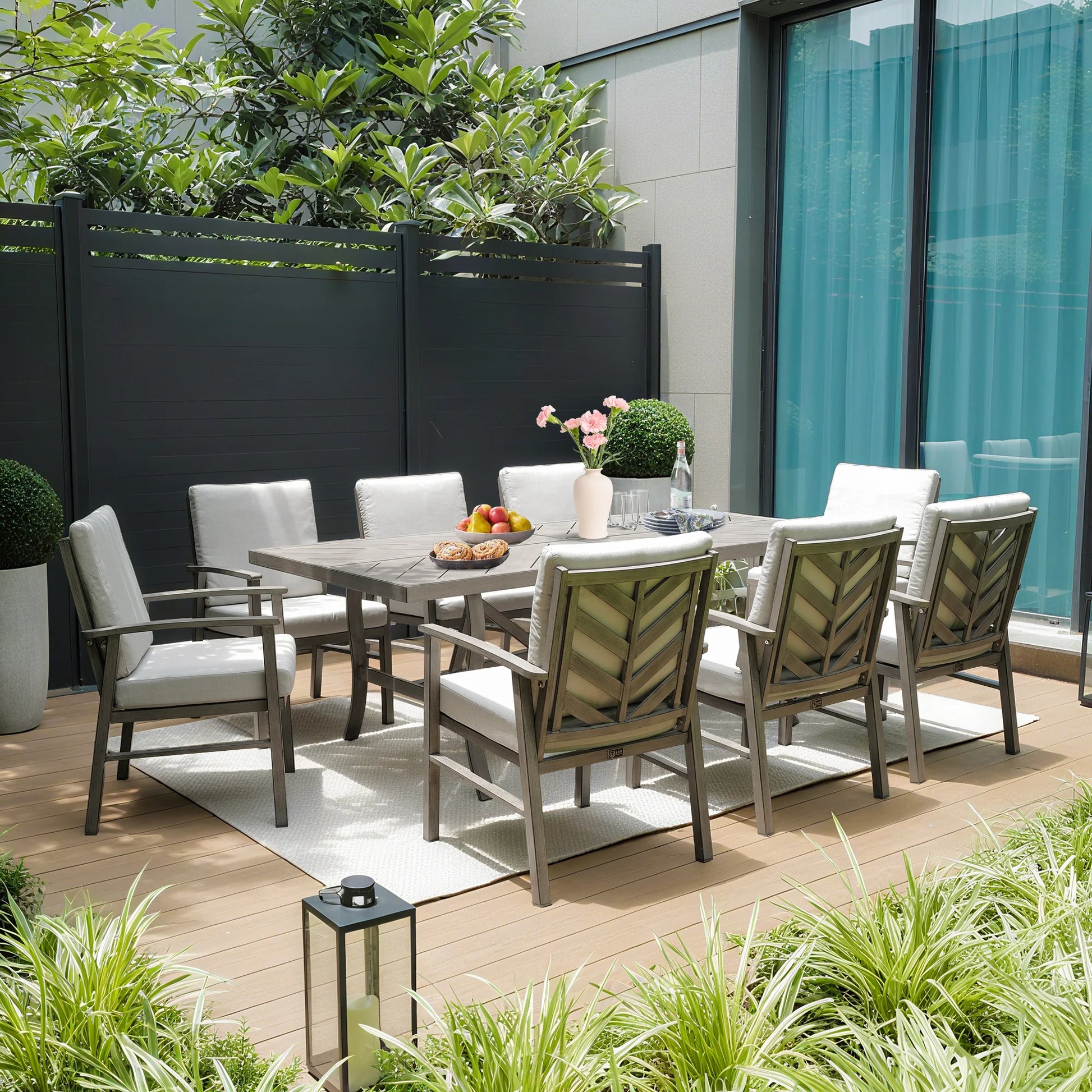 Outdoor dining set with table and chairs on a wooden deck, surrounded by plants and a building.