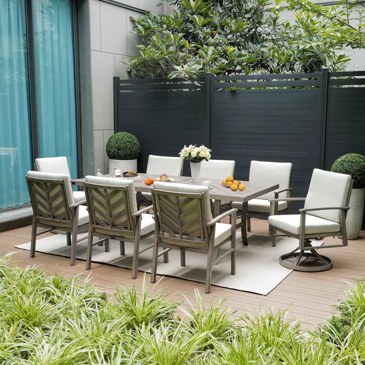 Outdoor patio set with table and chairs on a wooden deck, surrounded by plants and a building.
