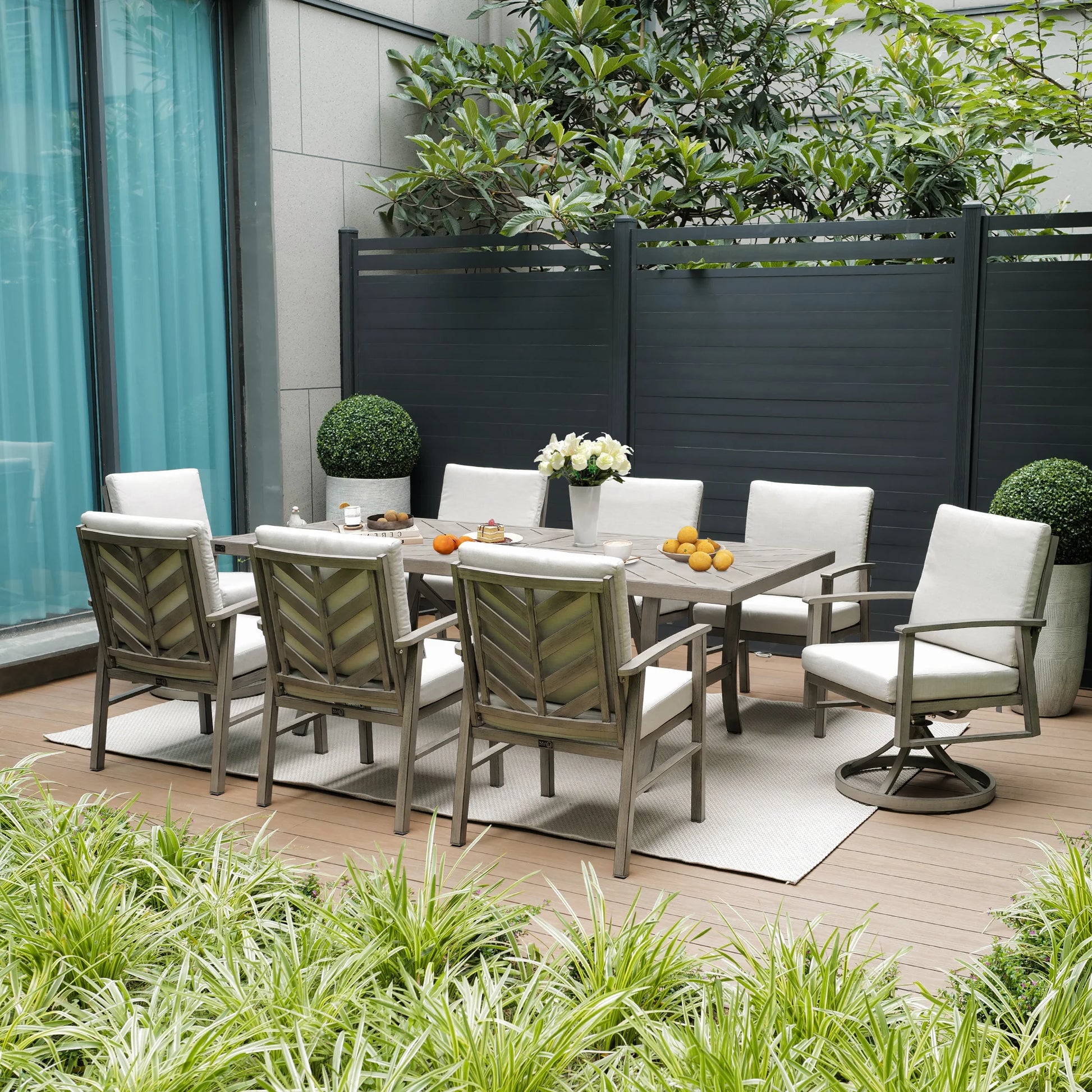Outdoor patio set with table and chairs on a wooden deck, surrounded by plants and a building.