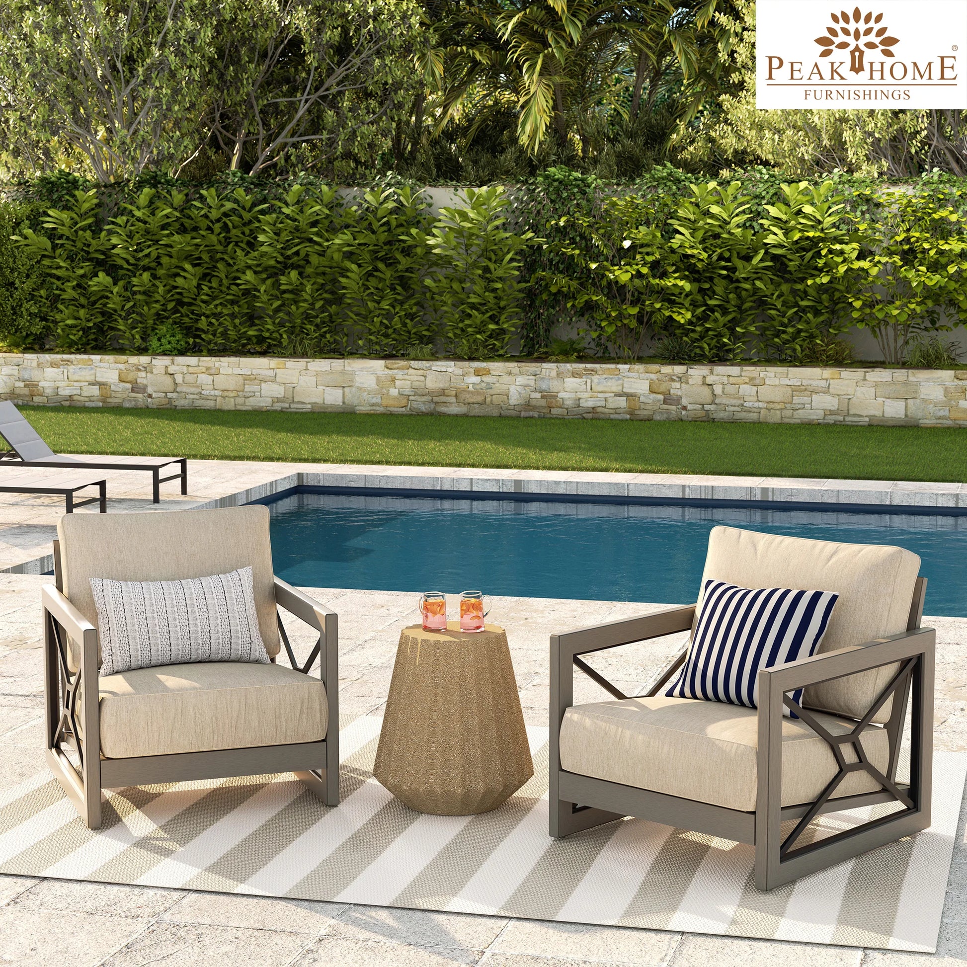 Two beige patio aluminum club chairs with sunbrella cushions placed on a tiled floor next to a round wooden side table with items on it, with a pool and greenery in the background.