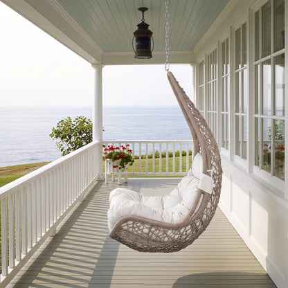 Side angle lifestyle view of Peak Home Furnishings teardrop rattan patio swing chair hanging from a porch ceiling overlooking the ocean.