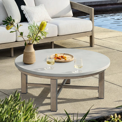 Round outdoor coffee table with a vase of flowers and a plate of fruit on a patio by a pool.