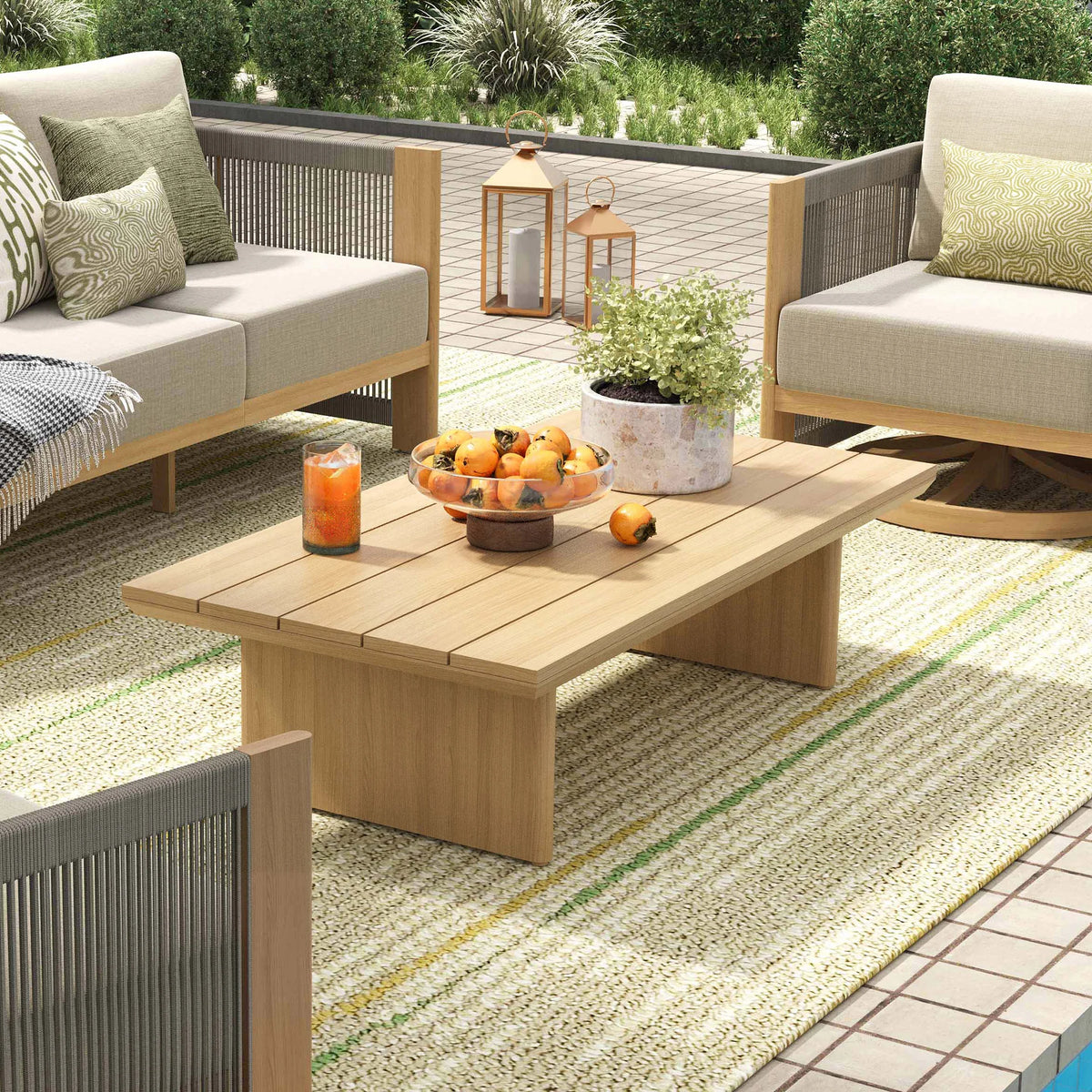 Modern rectangular outdoor coffee table with a warm teak finish surface styled with a bowl of fruit next to a poolside lounge area.