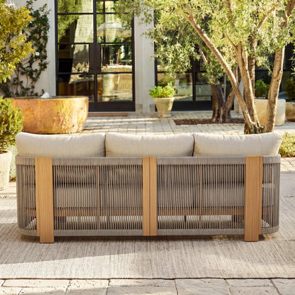 Outdoor sofa with beige cushions on a patio area with plants and a building in the background