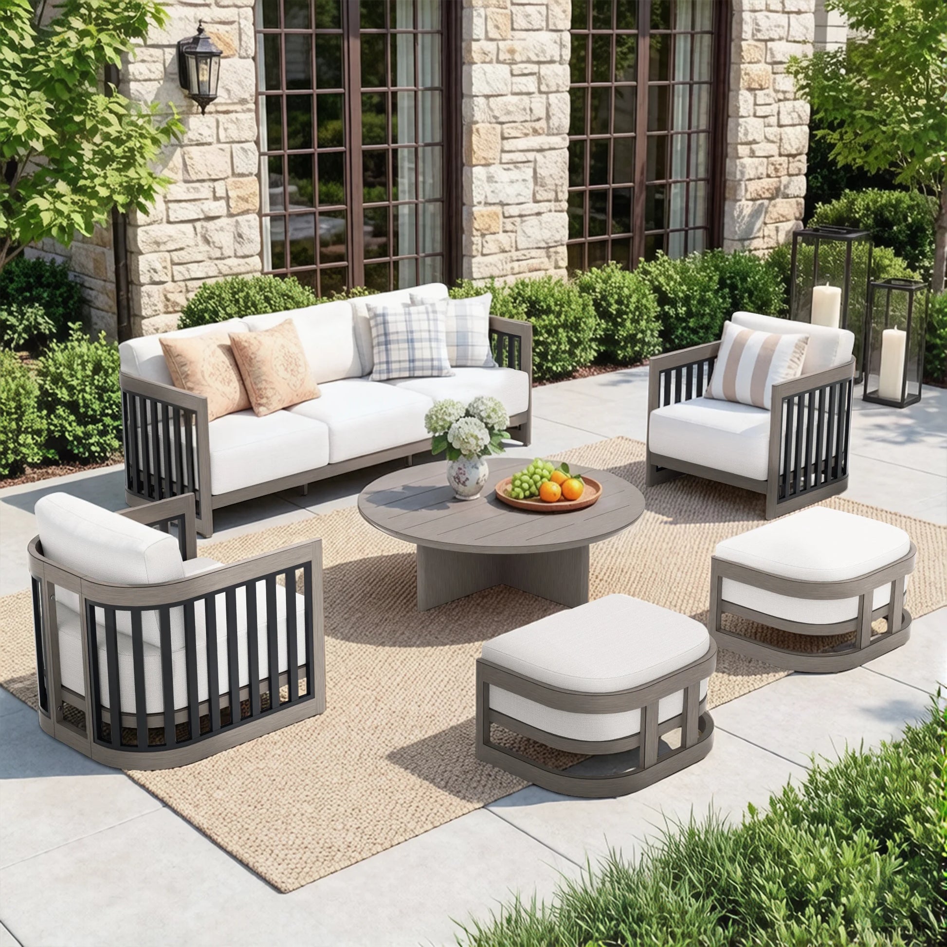 Outdoor patio furniture set including a sofa, two armchairs, two ottomans, and a round coffee table on a woven rug in front of a stone house.