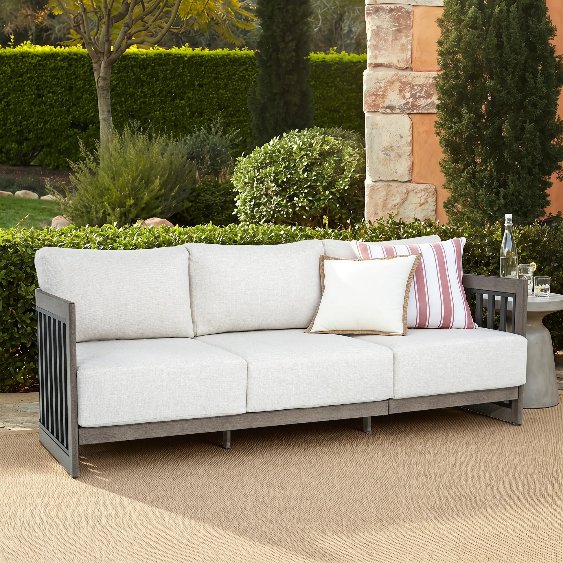 Outdoor patio sofa with light beige cushions and a dark gray frame, styled with two accent pillows in a garden setting.