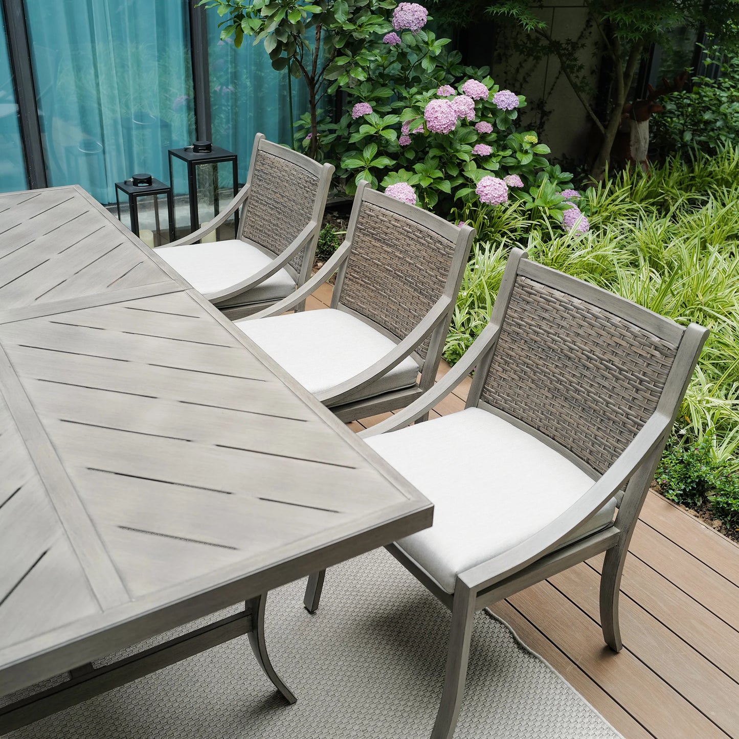 Outdoor patio set with a table and chairs on a wooden deck with plants in the background.