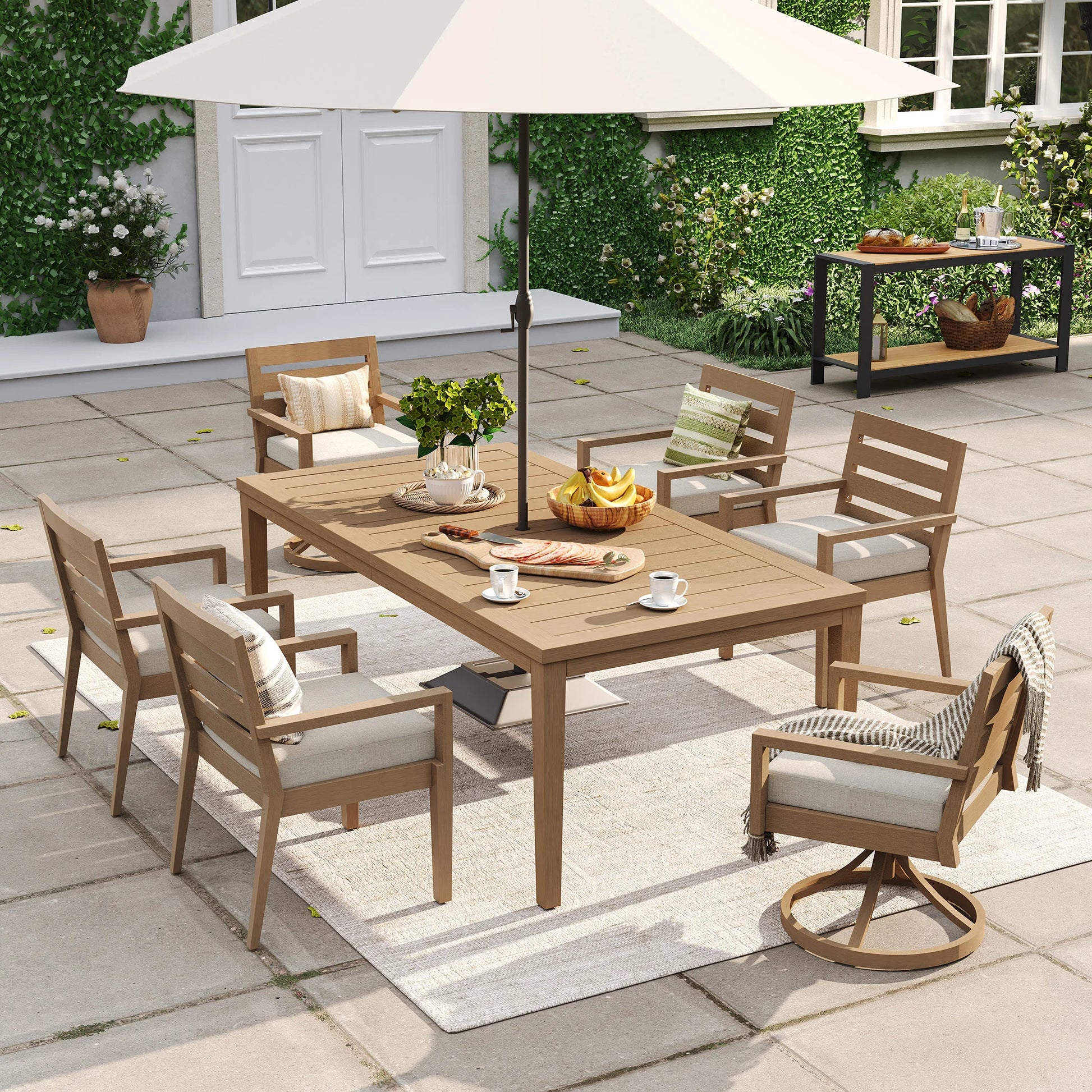 Alcudia 7-piece outdoor dining set featuring an 83-inch faux oak table with a large white patio umbrella, paired with 4 stationary chairs and 2 swivel rockers on a backyard stone terrace.