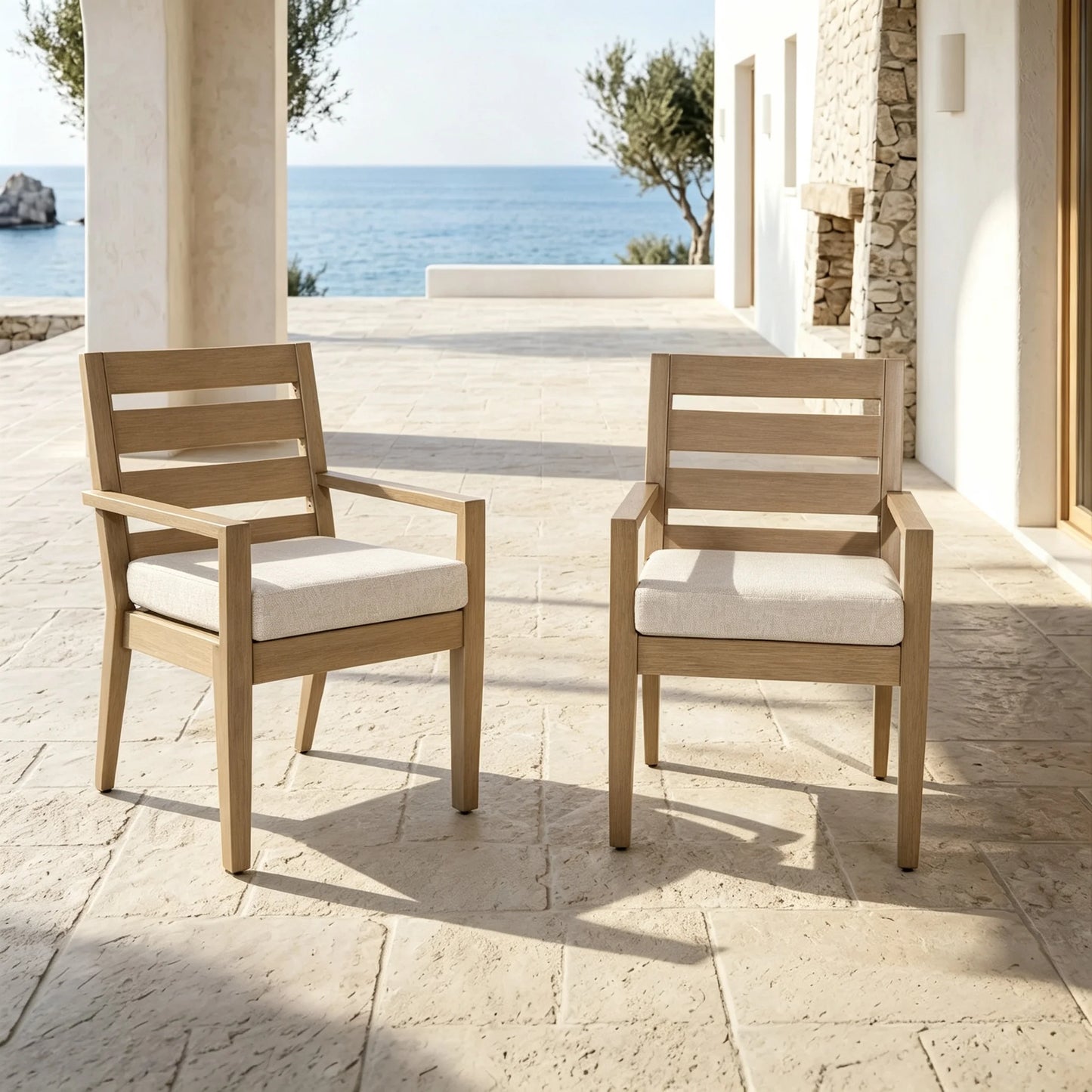 A pair of Alcudia faux wood aluminum patio dining armchairs with beige cushions styled on a luxury villa terrace overlooking the sea.