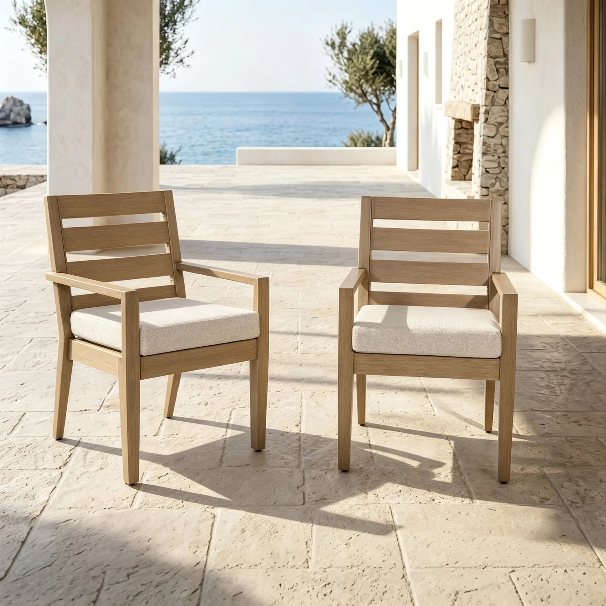 A pair of Alcudia faux wood aluminum patio dining armchairs with beige cushions styled on a luxury villa terrace overlooking the sea.