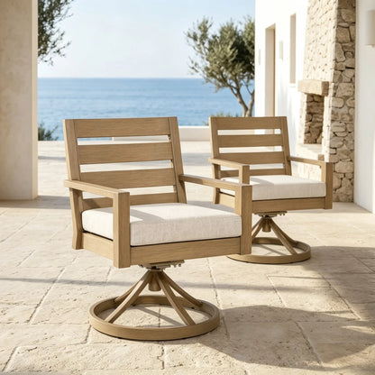 A pair of Alcudia luxury swivel patio armchairs with woven beige cushions arranged on a mediterranean-style stone terrace with a blue ocean view.