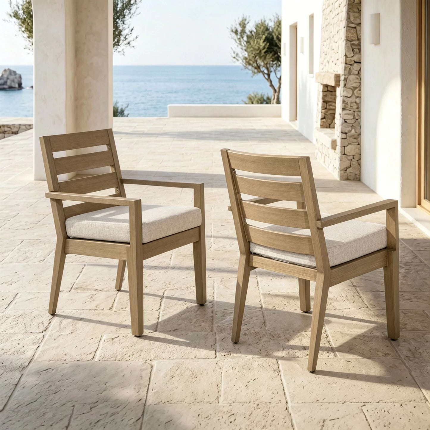 Front and back angles of two Alcudia outdoor dining armchairs featuring slatted backrests and hand-brushed wood finish with a coastal backdrop.