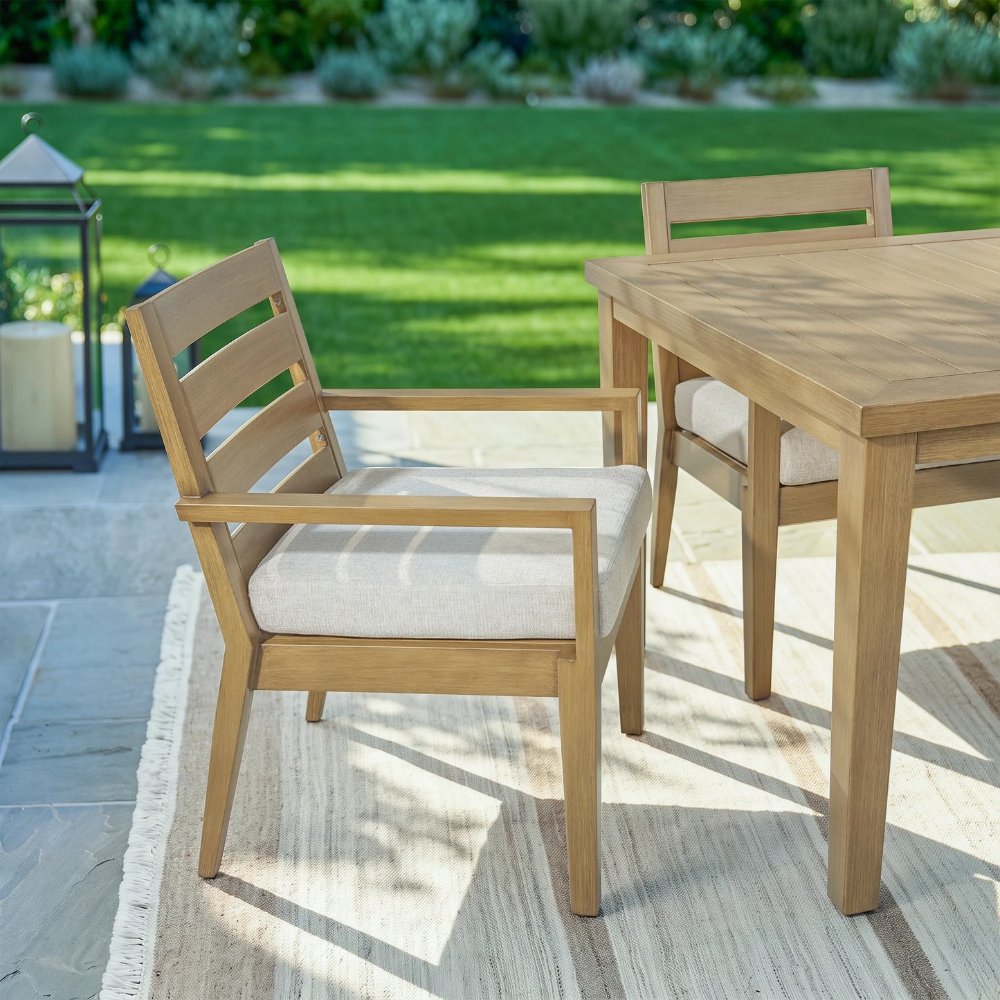 Angled view of an Alcudia faux oak aluminum outdoor dining chair with a beige cushion, styled next to a matching table on a patterned patio rug with a green lawn background.