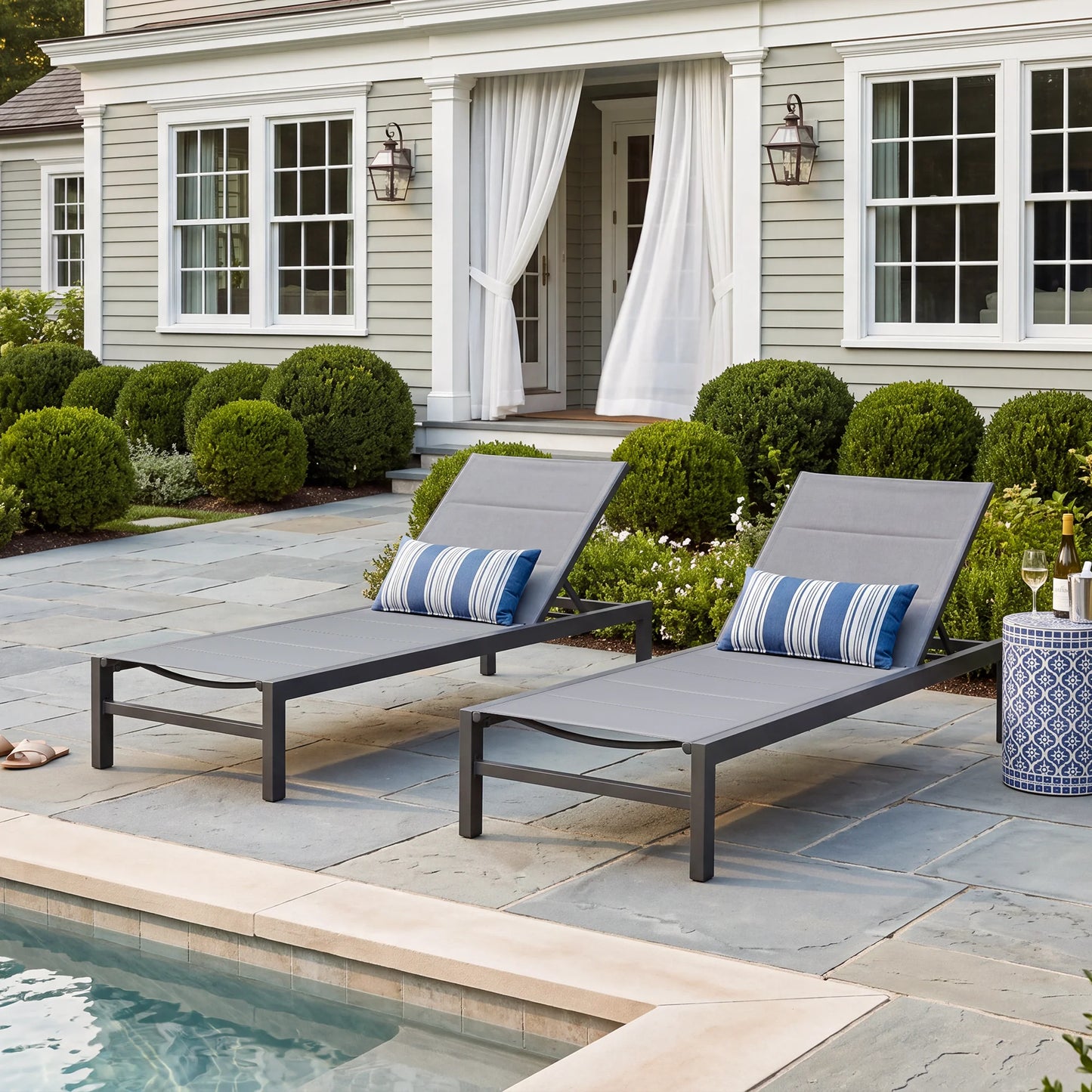 Two dark gray powder-coated aluminum chaise lounge chairs with padded charcoal sling fabric and blue striped lumbar pillows on a stone patio by a swimming pool.