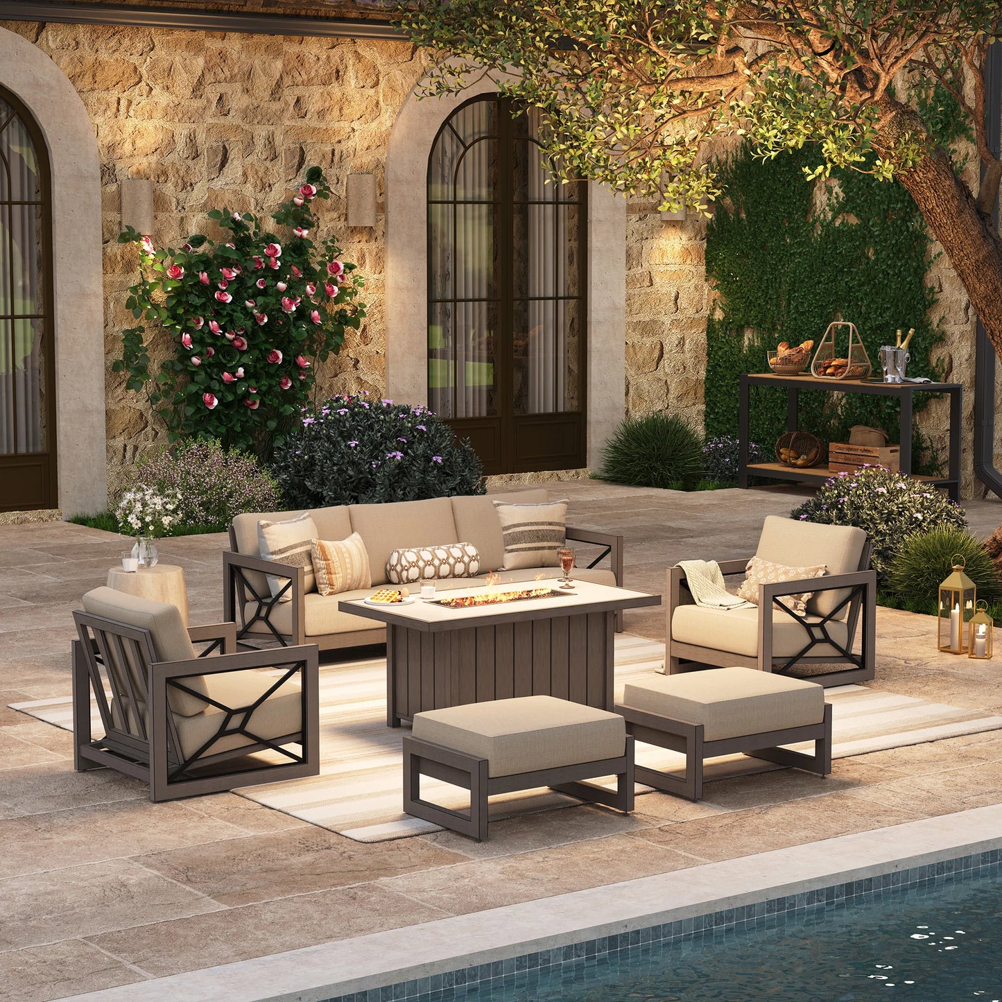 Upscale outdoor entertaining space at night featuring the Marindo 6-piece aluminum seating set, with amber flames glowing from the stainless steel fire pit table surrounded by a sofa, club chairs, and ottomans.