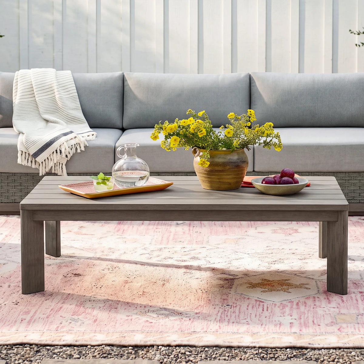 Front view of the modern Manarola grey slatted patio coffee table decorated with a flower vase and pitcher in front of an outdoor sofa.
