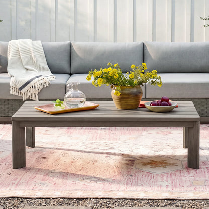 Front view of the modern Manarola grey slatted patio coffee table decorated with a flower vase and pitcher in front of an outdoor sofa.