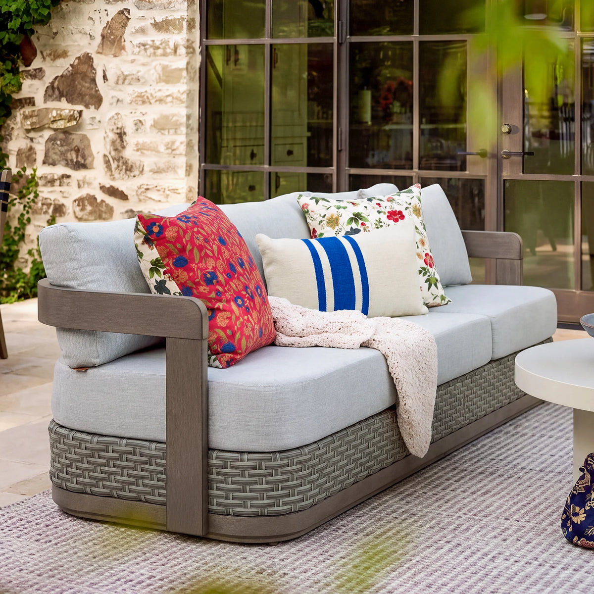 Side angle view of the Manarola modern 3-seater fixed outdoor couch styled with vibrant decorative pillows and a throw blanket on a chic patio rug.